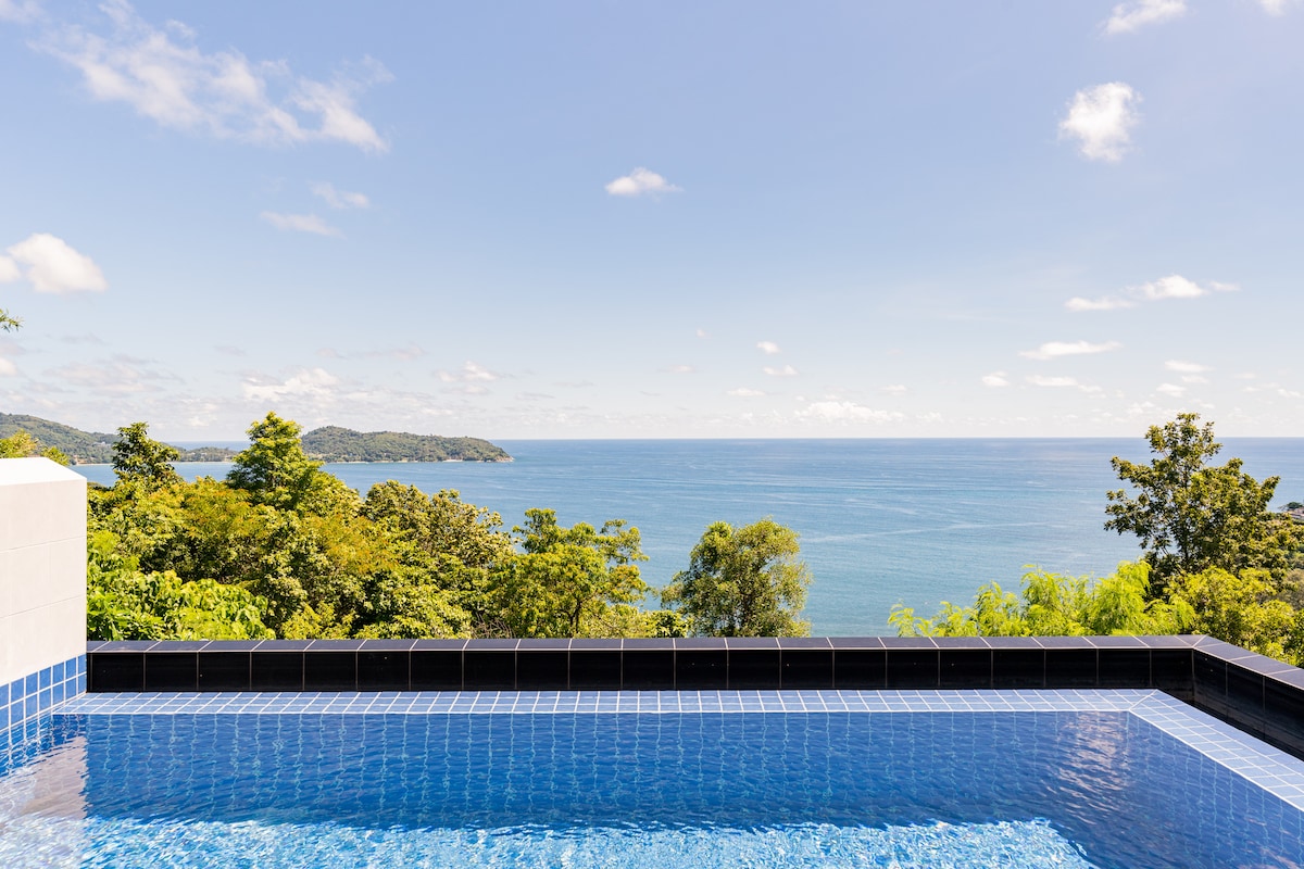 A private infinity pool reflects the vibrant blue sky and the surrounding greenery. The calm sea stretches out in the distance, offering a panoramic view of the horizon where water meets land.