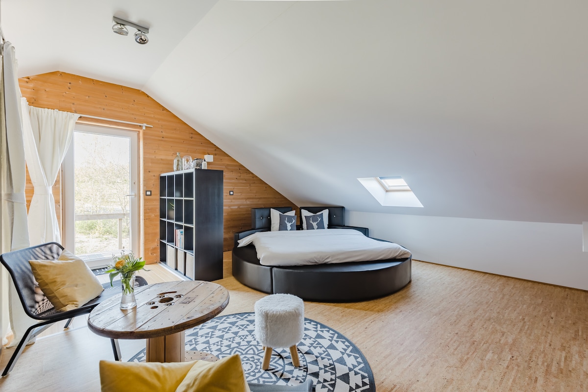 An airy bedroom features a circular bed with neutral bedding against a wooden panel wall. A small round table is positioned nearby, accompanied by a chair and a cozy rug. Natural light enters through a slanted window and a door leading to a balcony.