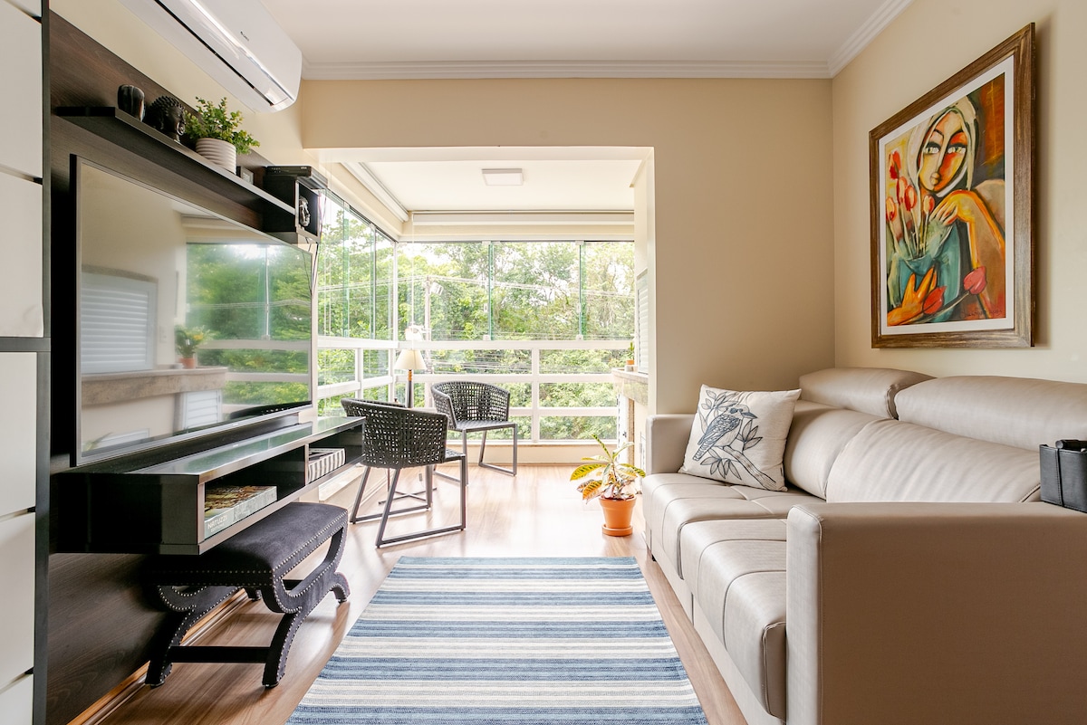 A light-filled living area features a comfortable beige sofa positioned across from a sleek black entertainment unit. A striped rug adds a touch of color, while a balcony view is framed by glass walls, showcasing greenery outdoors. A potted plant brings a natural element to the space.