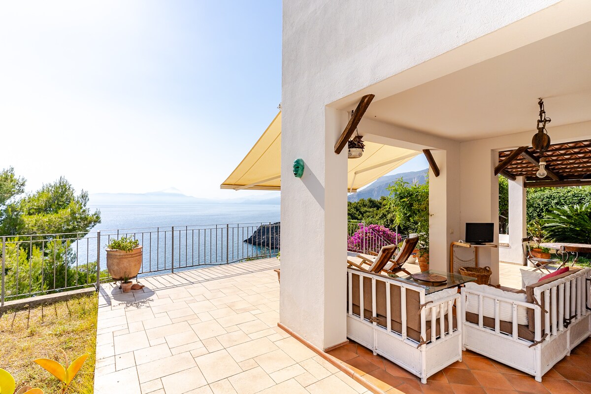 A spacious terrace overlooks the tranquil waters of the Gulf of Policastro. Comfortable seating arrangements are placed under a shaded area, complemented by lush greenery and vibrant natural views in the background. Sunlight reflects off the serene sea, enhancing the relaxing ambiance.