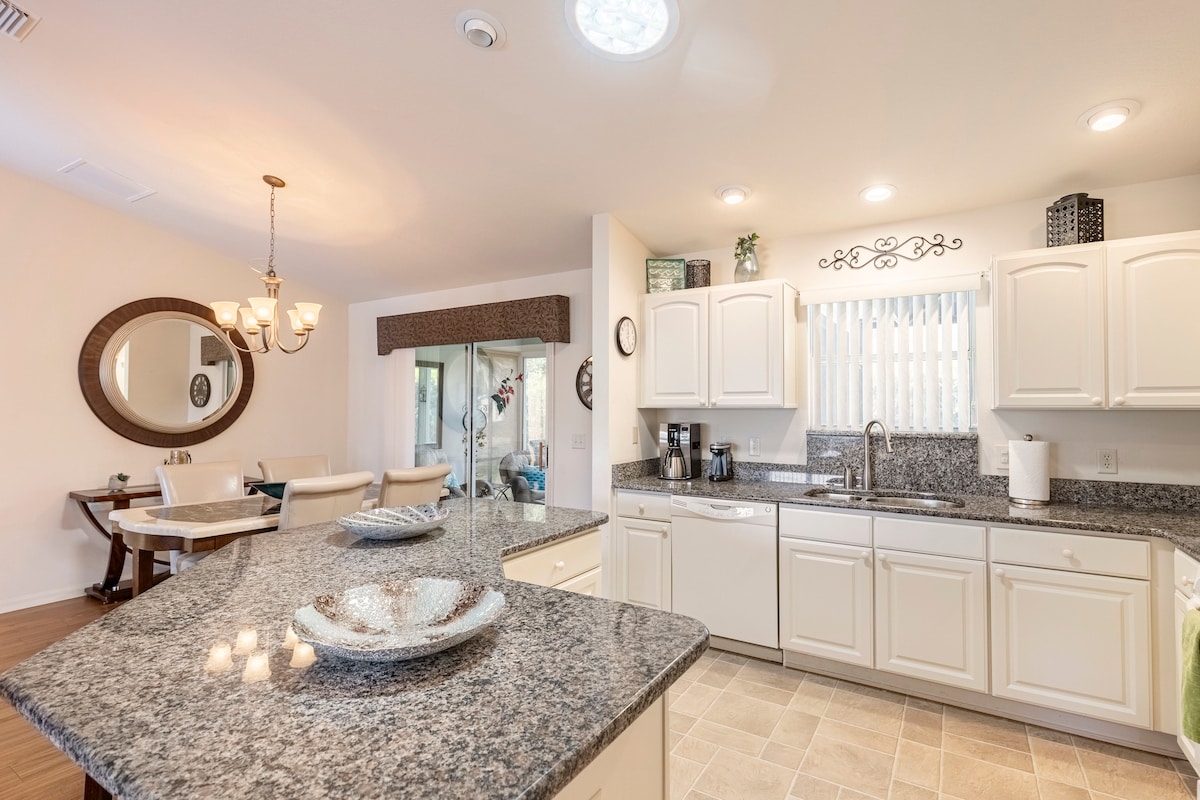 A modern kitchen is featured with granite countertops, white cabinetry, and stainless steel appliances. A spacious island offers additional counter space. A dining area with a circular mirror and light fixture is visible in the background, providing a welcoming atmosphere for meals.