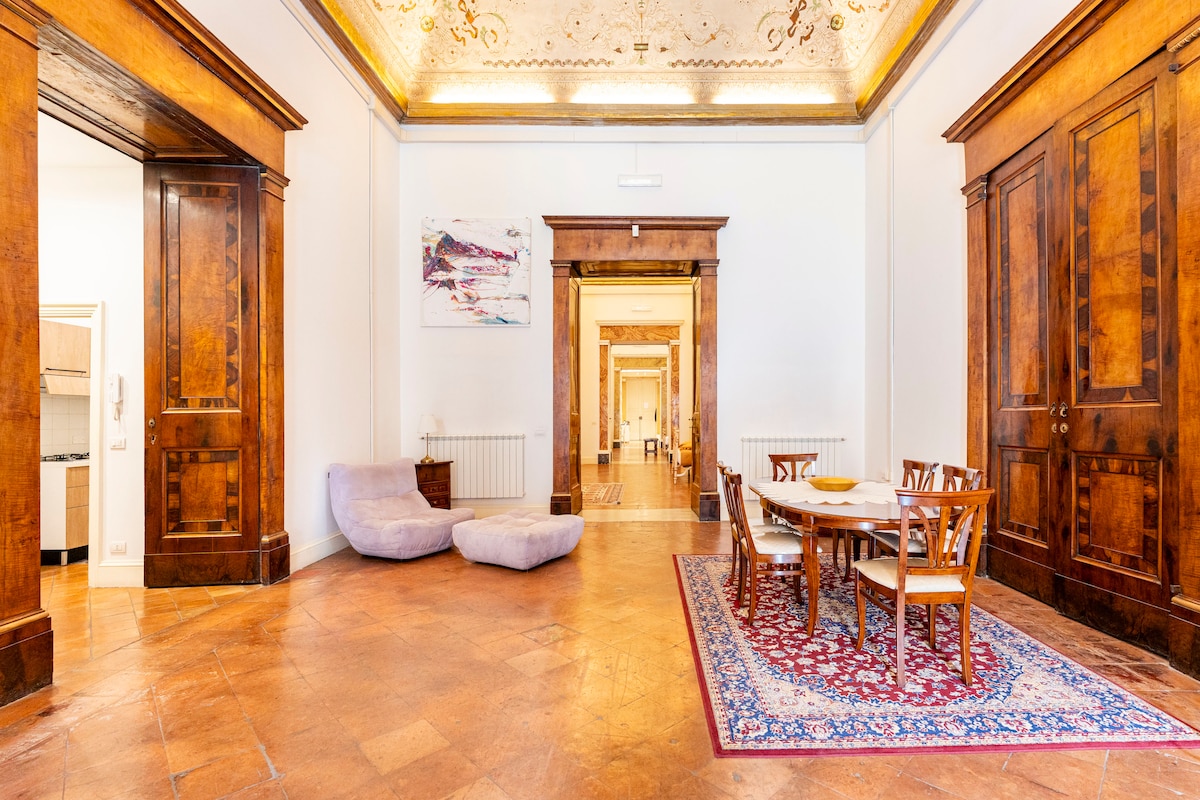A spacious living area features high ceilings adorned with intricate frescos. Elegant wooden cabinets frame the room, while a round dining table accommodates several chairs. A plush seating area is visible in the background, enhancing the room's inviting character and charm.