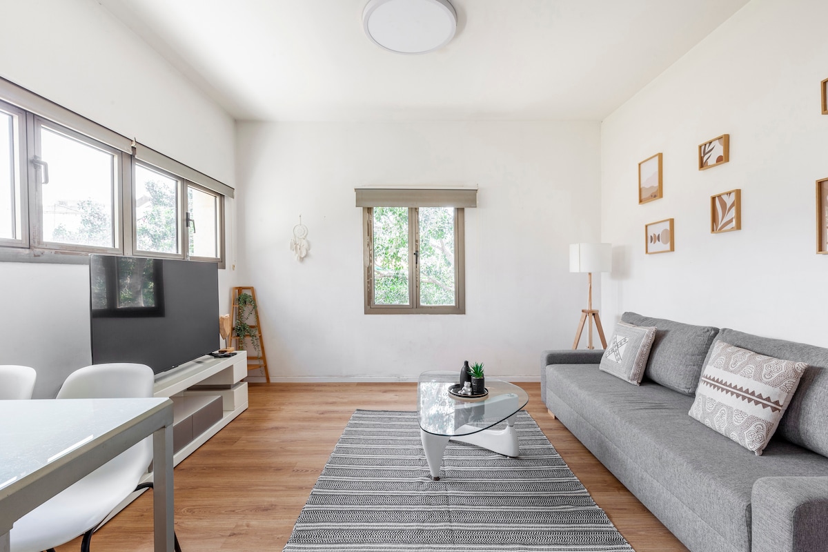 A bright living area is featured, showcasing a gray sofa paired with decorative pillows. Natural light enters through two windows framed by light wood. A glass coffee table sits on a striped area rug, with a small plant as a centerpiece. Minimalist wall art is displayed.