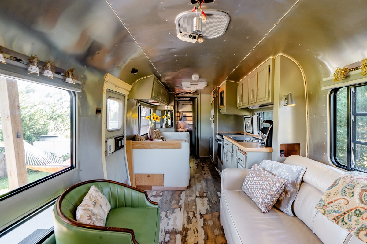 An inviting interior features a spacious living area with a comfortable light-colored sofa and a green armchair. Natural light floods through large windows, highlighting the rustic wooden flooring. Kitchenette appliances are visible, along with cabinets and decor accents that create a cozy atmosphere.