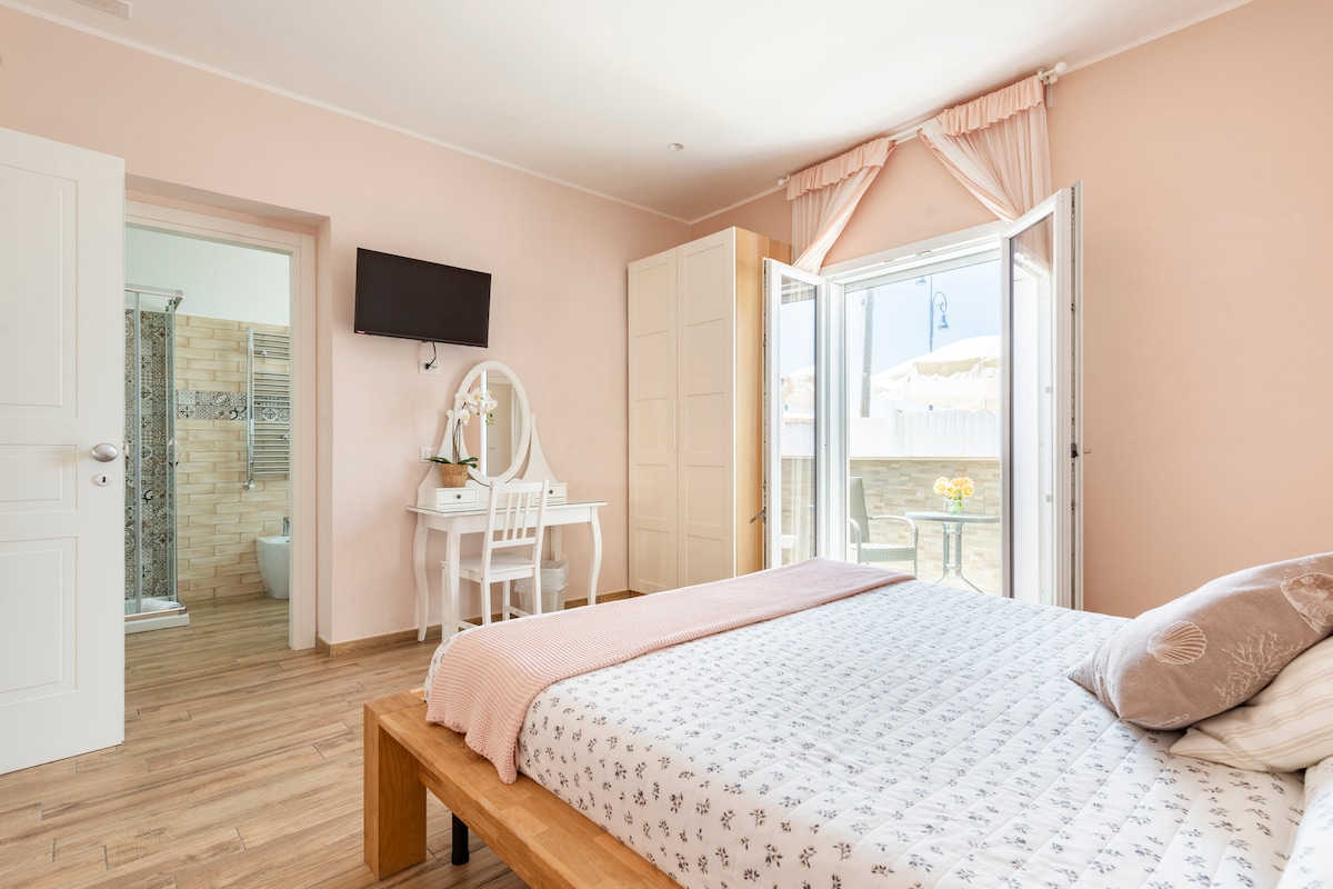 A serene bedroom is featured with soft pink walls and a wooden bedframe. Natural light fills the space through open doors leading to an outdoor area. A small vanity with a chair and a television on the wall are visible, along with an ensuite bathroom entrance.