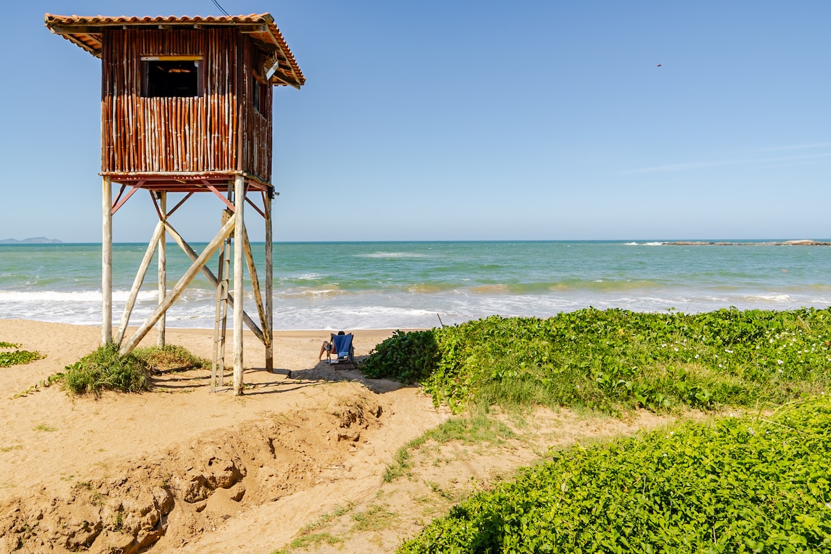 A wooden lookout tower stands on stilts along a sandy beach, providing views of the ocean. Two chairs are positioned on the sand, facing the water, surrounded by lush green vegetation. The sky above is clear, with gentle waves lapping at the shoreline.