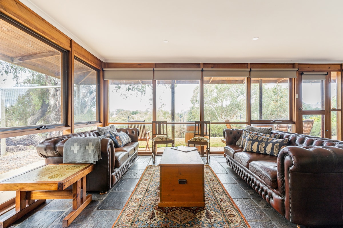A comfortable living area features dark leather sofas, a wooden coffee table, and an intricate area rug. Large windows provide a panoramic view of the surrounding landscape, allowing natural light to fill the space. Stylish wooden chairs are positioned at one end, inviting relaxation.