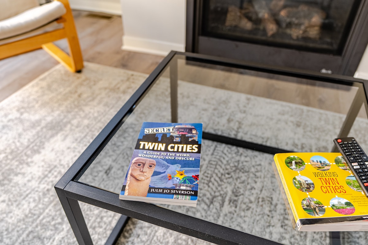 A glass-topped coffee table is featured in the living area, displaying a travel book titled 'Secret Twin Cities' and a guidebook to 'Walking Twin Cities.' A remote control rests beside the books, positioned on a light-colored area rug. The room is brightened by natural light from nearby windows.