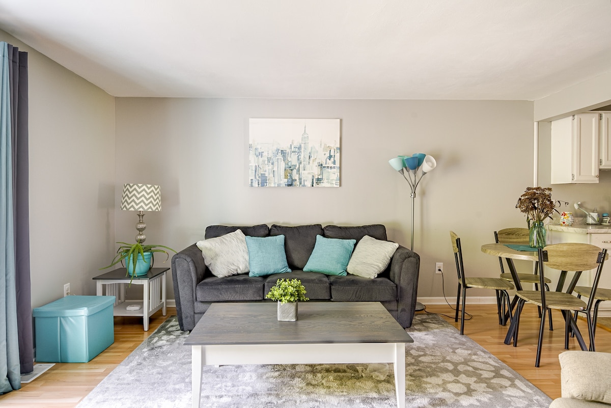 The living area features a comfortable, dark sectional sofa adorned with teal cushions, complemented by artwork on the wall. A light blue table lamp and potted plant add a touch of greenery. A dining table with chairs is visible in the background, alongside a well-lit kitchen space.