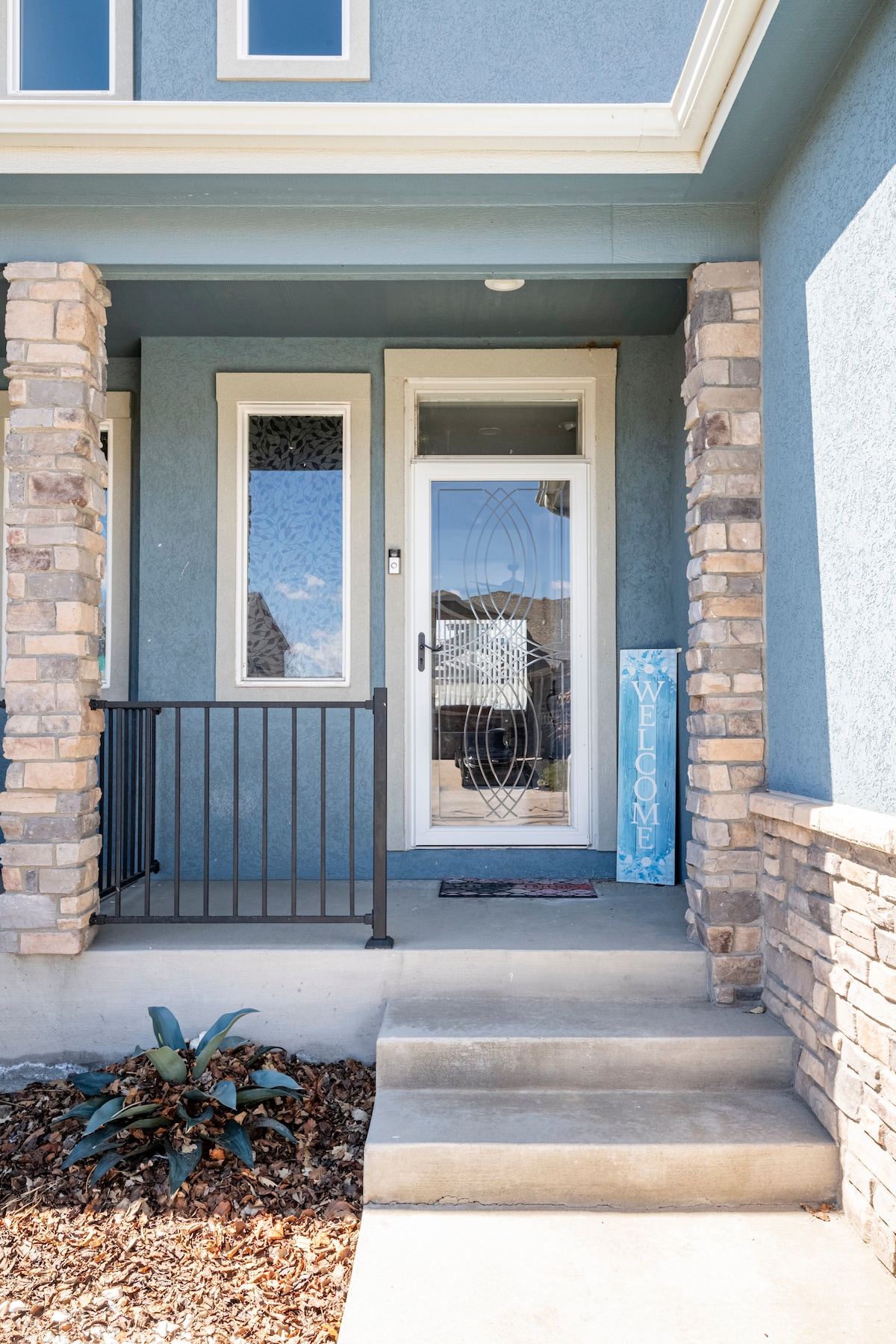 The entryway features a welcoming front door adorned with a decorative glass design. Flanked by stone pillars, a small step leads up to the entrance. A potted plant adds a touch of greenery beside the door, contributing to the inviting exterior.