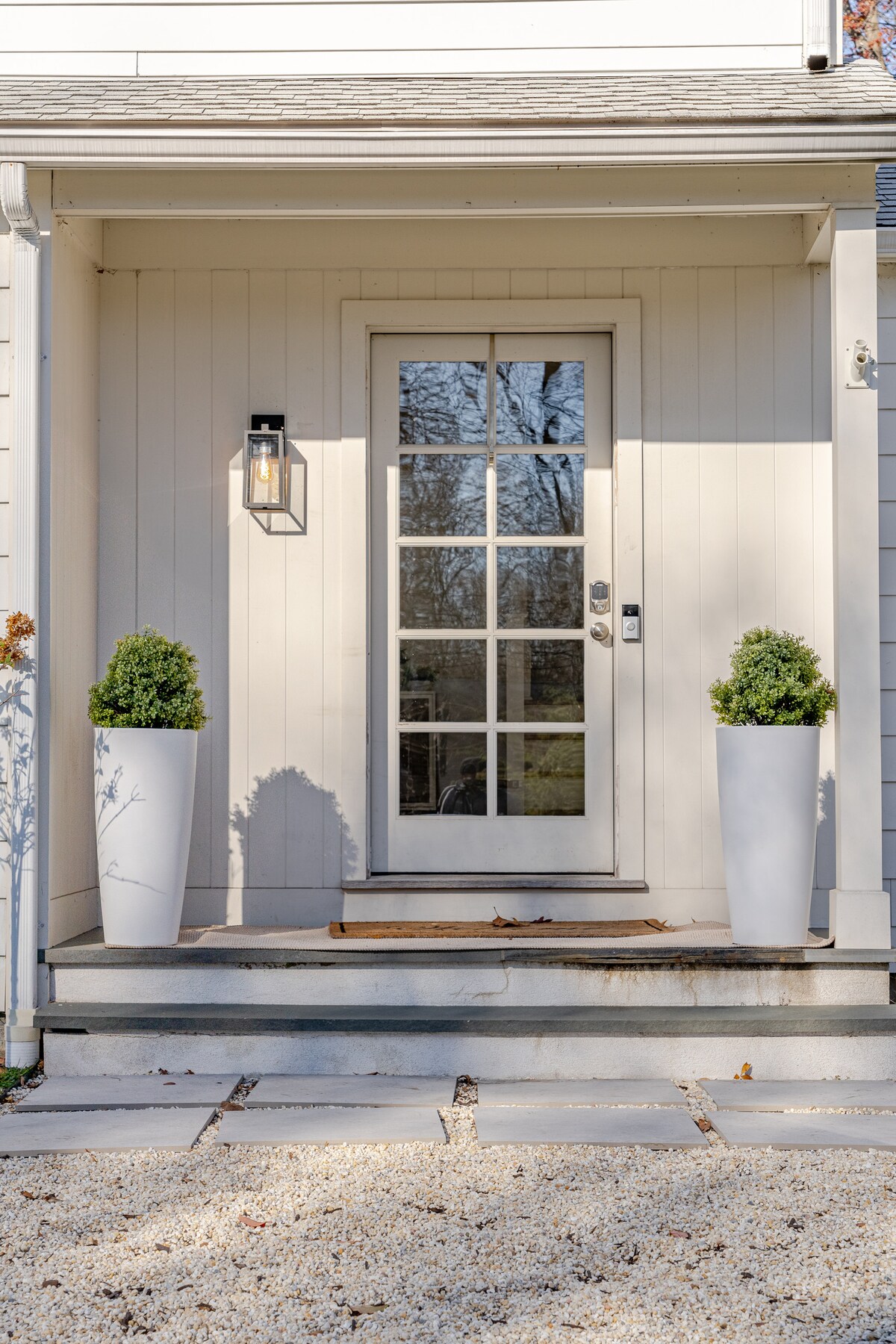 A welcoming entrance features a light-colored front door with multiple glass panes, framed by tall white planters containing neatly trimmed greenery. A small welcome mat lies on the porch, and a stone pathway leads towards the door, enhancing accessibility.