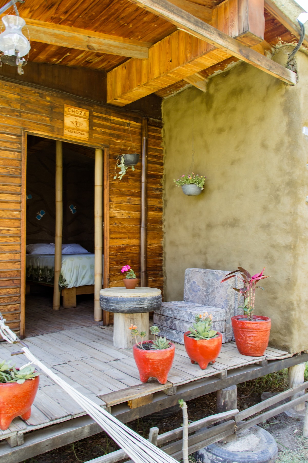 A welcoming terrace leads to the entrance of the rustic dwelling, featuring wooden walls and a clay exterior. Potted plants add greenery, while a small table and comfortable seating offer a relaxing space. Two hanging lights are placed above, contributing to the inviting ambiance.
