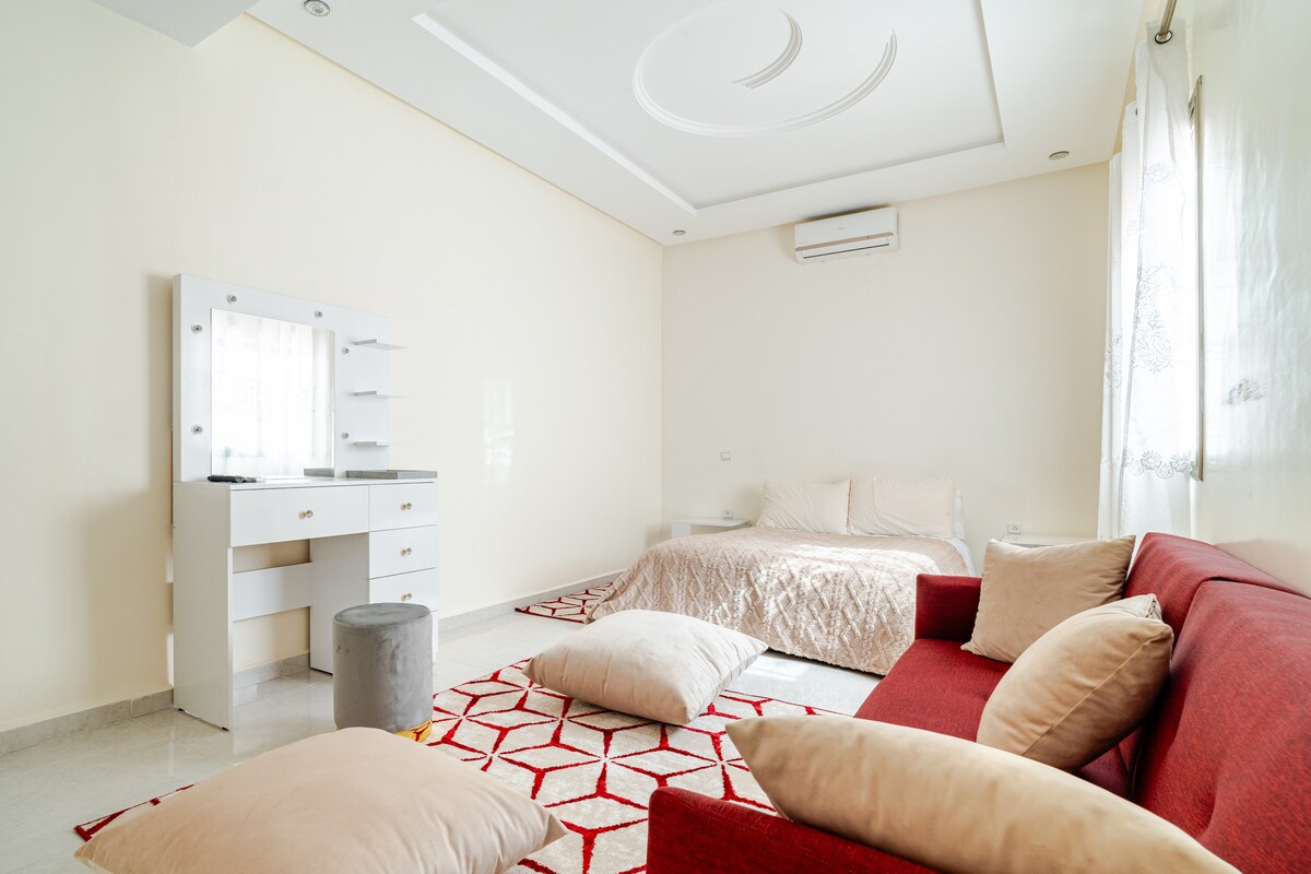 A bright and spacious bedroom features a large bed and a red sofa, creating a comfortable setting. Soft pillows are arranged on a geometric-patterned rug. Natural light enters through sheer curtains, highlighting the room's neutral walls and ceiling design.