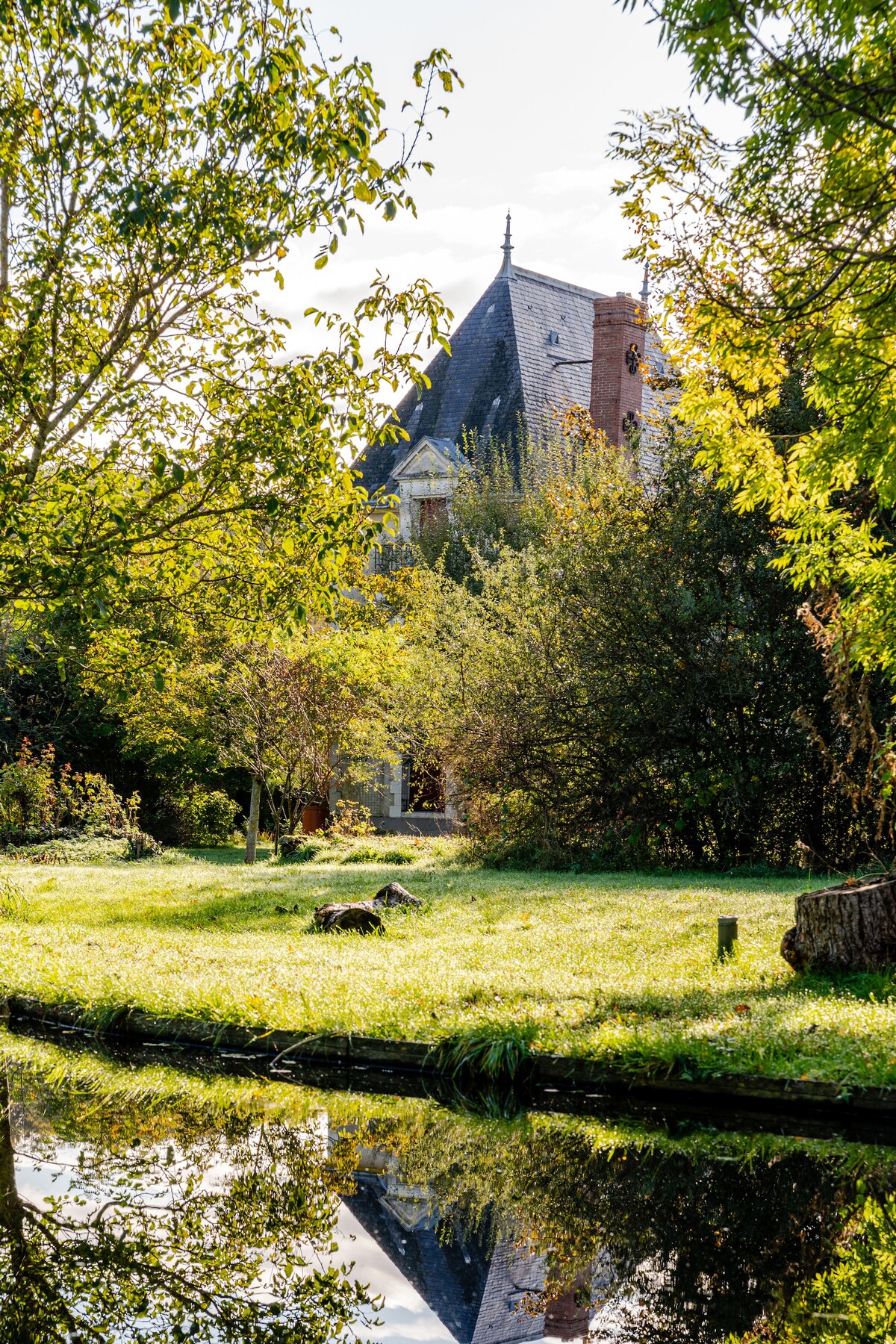 The exterior view of La Combe is surrounded by lush greenery, featuring trees and shrubs that frame the charming structure. A serene pond reflects the building and the surrounding landscape, creating a peaceful ambiance in this private domain.