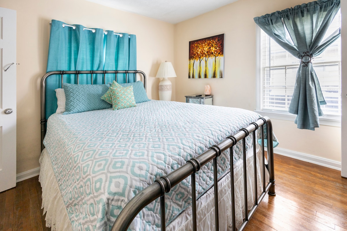 A spacious bed with a decorative quilt and accent pillows is positioned against a light-colored wall. Colorful artwork hangs above the bed, and natural light filters through a window adorned with sheer curtains. A bedside lamp and a white curtain frame another window.