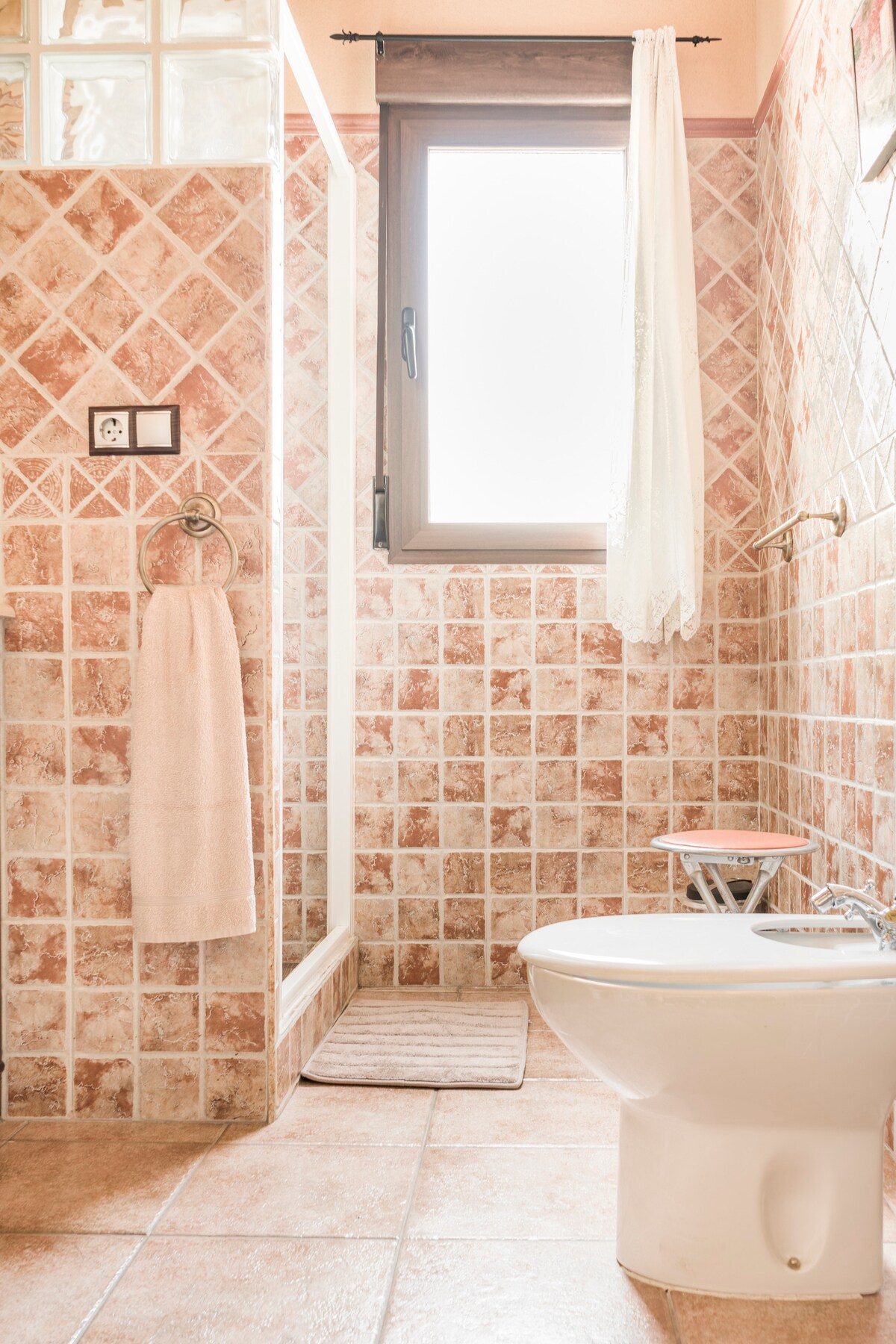A well-appointed bathroom features decorative tile patterns in soft earth tones. A shower area is enclosed by glass, with a plush mat in front. A white towel hangs gracefully on a ring, and a small window allows natural light to filter in, brightening the space.