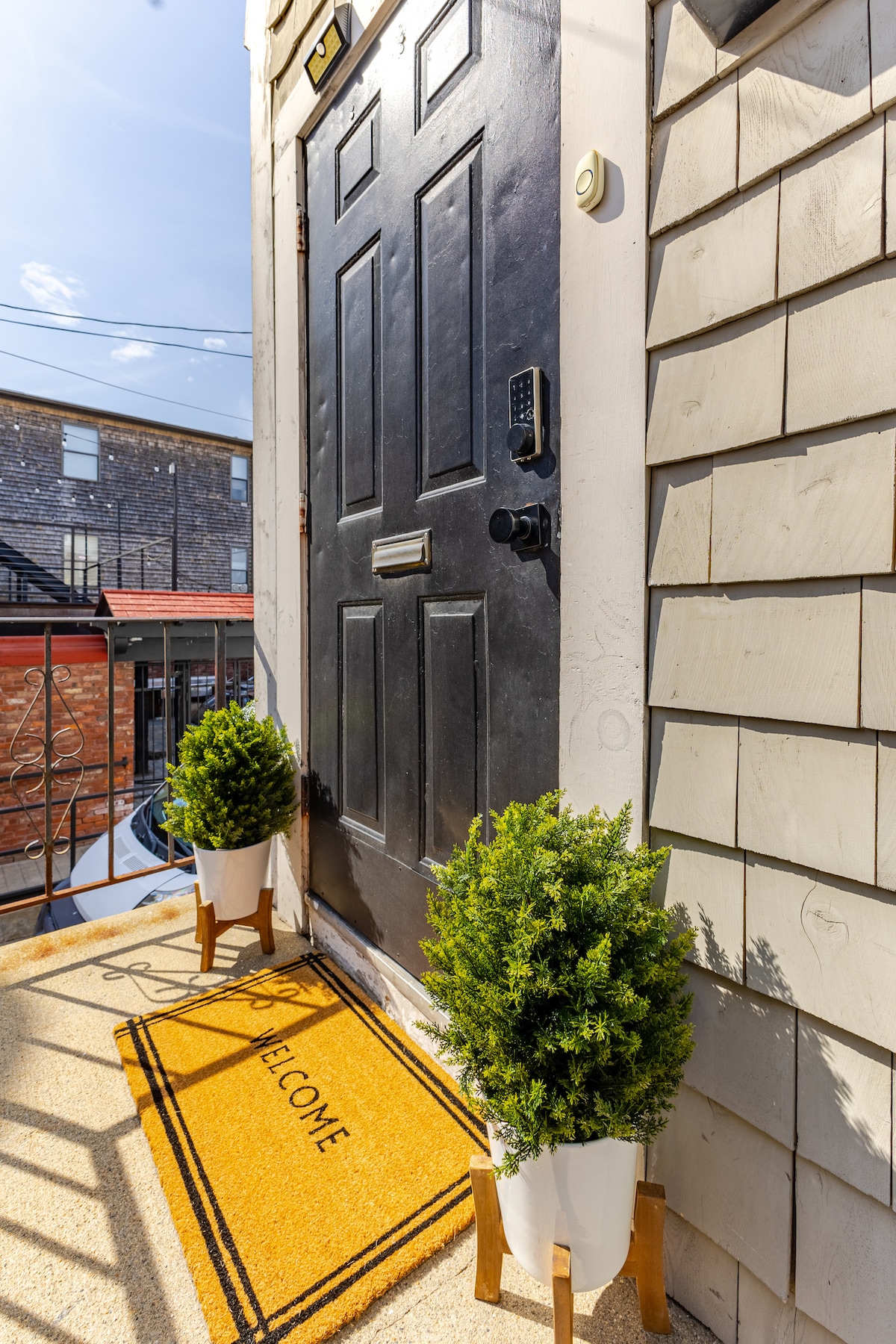 A welcoming entryway is visible with a black door featuring a keypad lock. A bright yellow doormat with the word 'Welcome' is placed in front, flanked by two small potted plants on wooden stands, bringing a touch of greenery to the space.