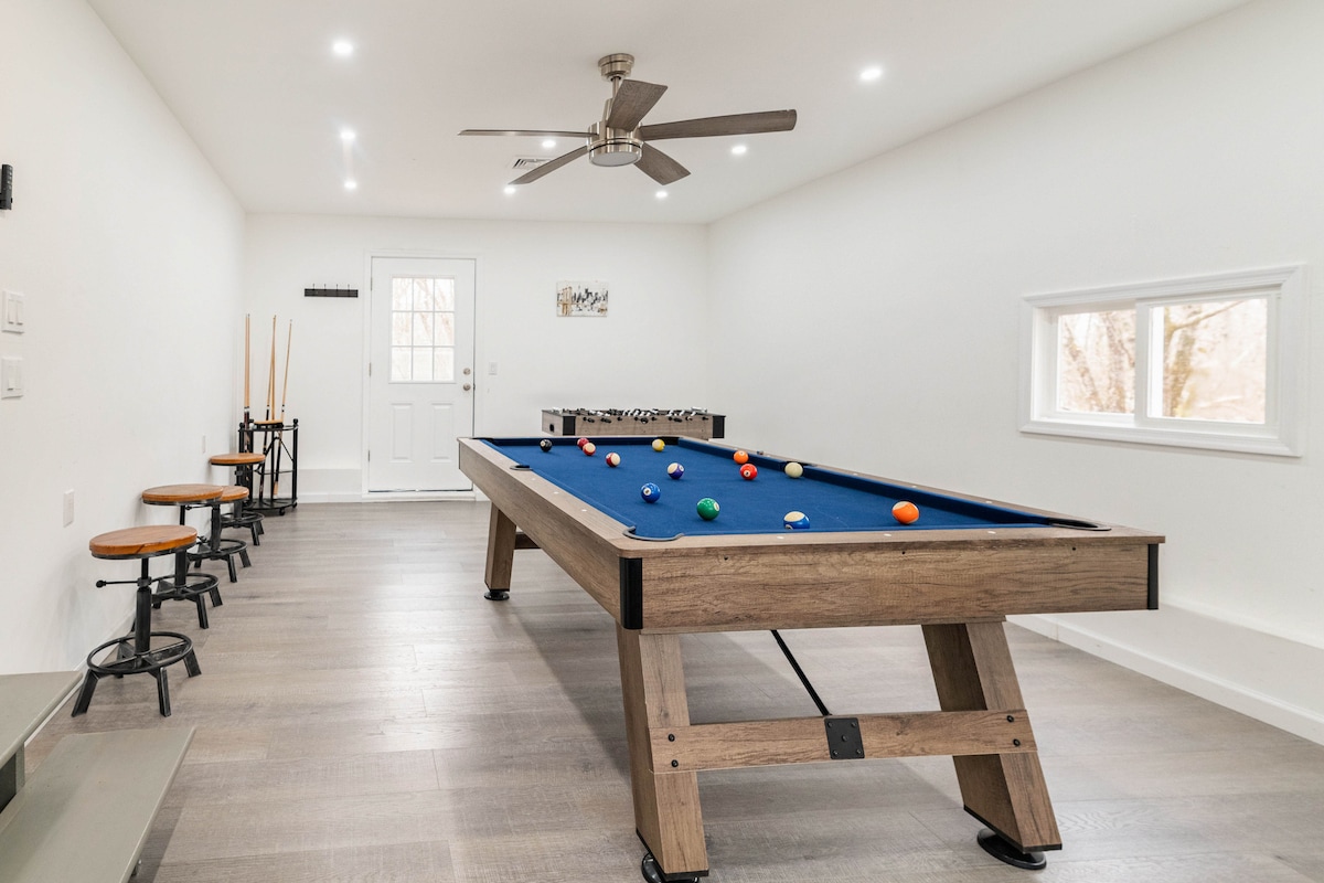 A spacious game room features a billiard table with colorful balls arranged on top. A ceiling fan is visible, along with several seating stools against a wall. Natural light enters through a nearby window, illuminating the modern wooden accents and light-colored walls.