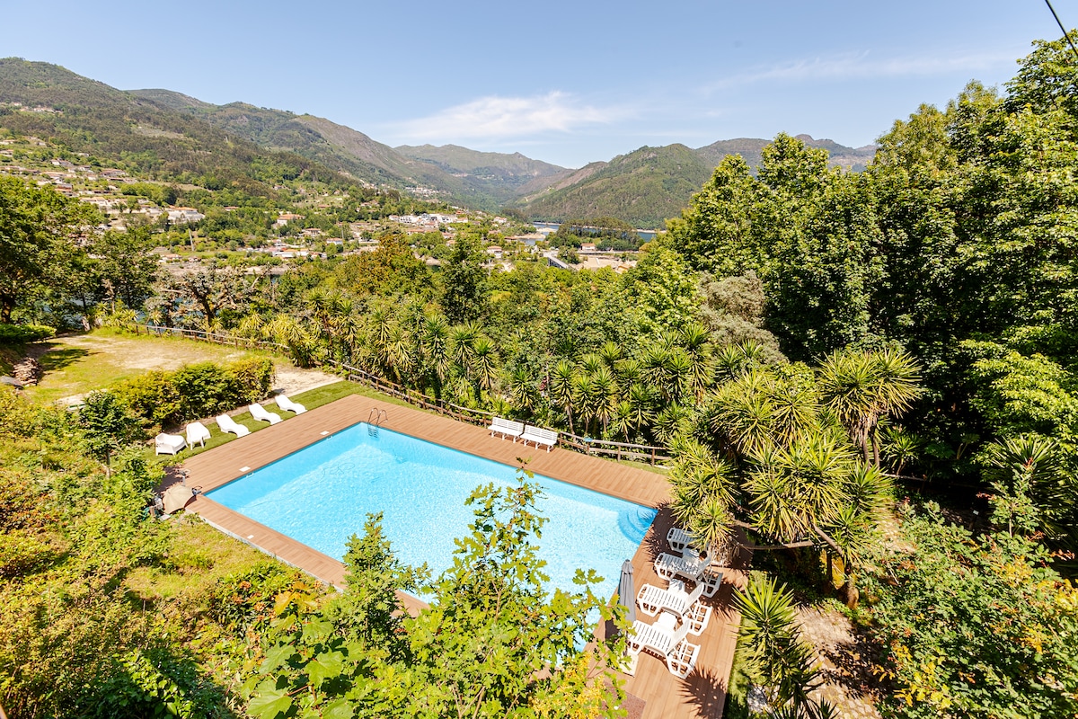 A spacious outdoor pool is surrounded by lush greenery and sun loungers. Scenic mountain views stretch in the background under a clear blue sky, creating a peaceful atmosphere for relaxation. The area is designed for enjoying the outdoors, with ample space for seating.