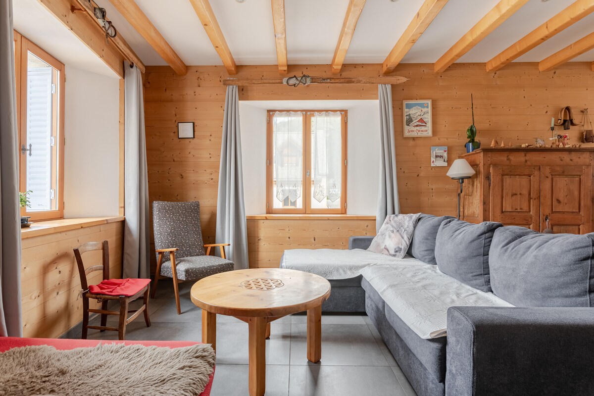 A spacious living area features warm wooden walls and exposed ceiling beams. A comfortable gray sofa is positioned around a round wooden coffee table. Two windows allow natural light to illuminate the space, complemented by a cozy armchair near the wall.