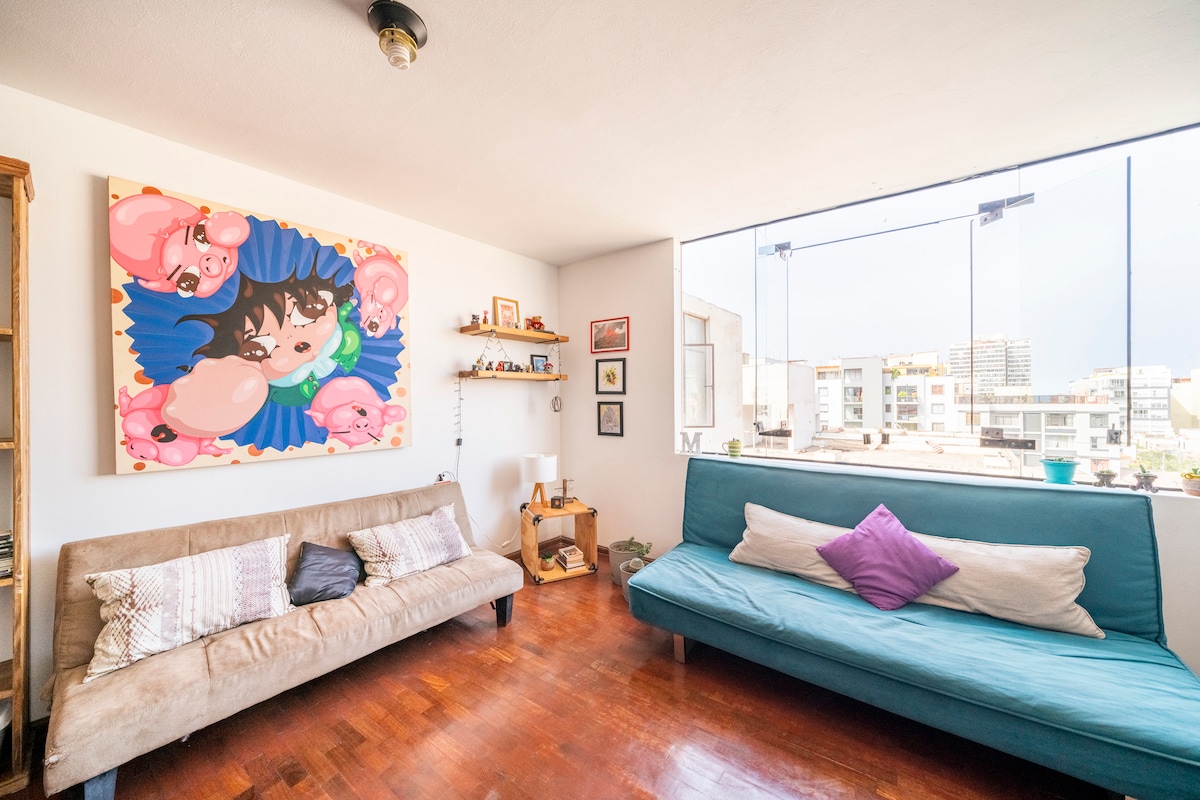 A bright and airy living space features two sofas, one adorned with decorative pillows. A colorful wall art piece draws attention, while large windows provide ample natural light and a view of the city. Shelves contain various decorative items.