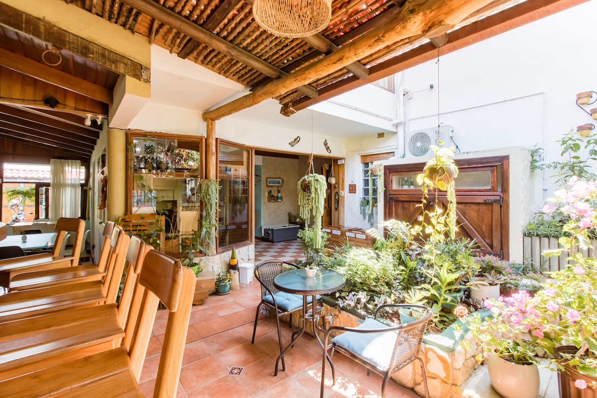 A serene courtyard features a table and seating surrounded by lush greenery and decorative plants. The area is illuminated by natural light and showcases wooden beams along the ceiling. Tall windows provide views into the garden, creating a peaceful atmosphere for relaxation.