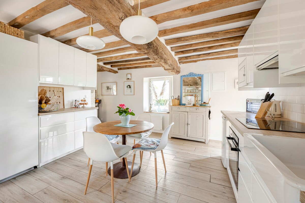 A modern kitchen features exposed wooden beams complemented by white cabinetry and a round wooden table surrounded by four chairs. Natural light enters through a window, illuminating the space where a floral arrangement sits centrally on the table.
