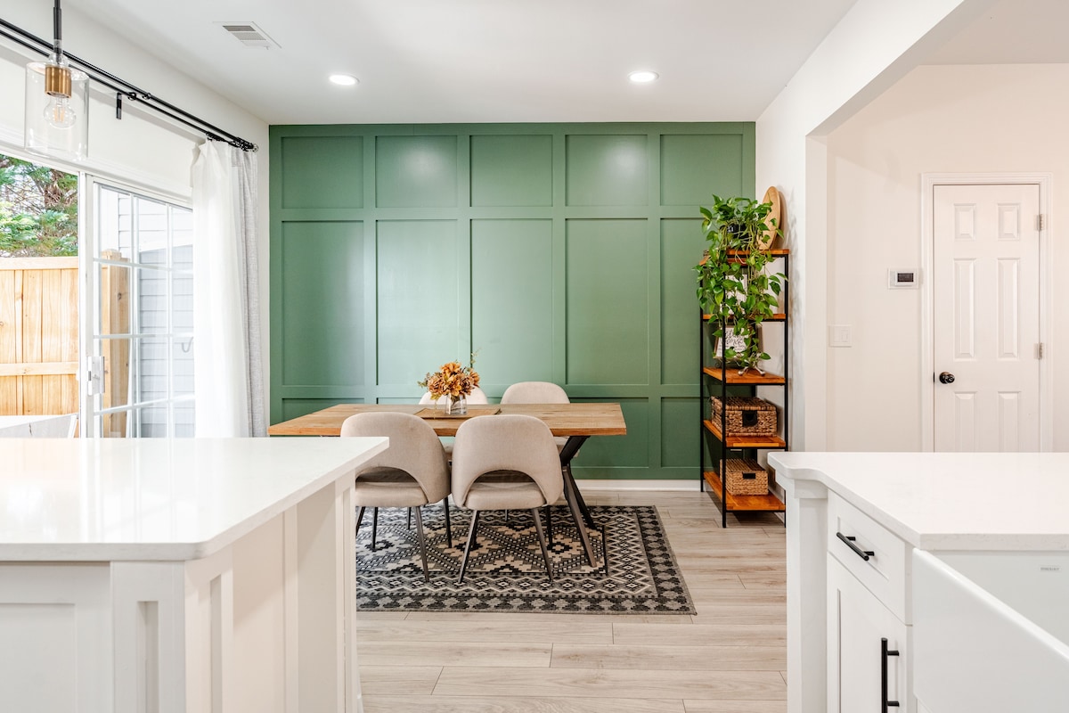 A dining area features a natural wood table surrounded by beige chairs, set against a green accent wall with geometric paneling. A decorative plant is placed on a shelf nearby, while light enters through sliding glass doors leading to an outdoor space.
