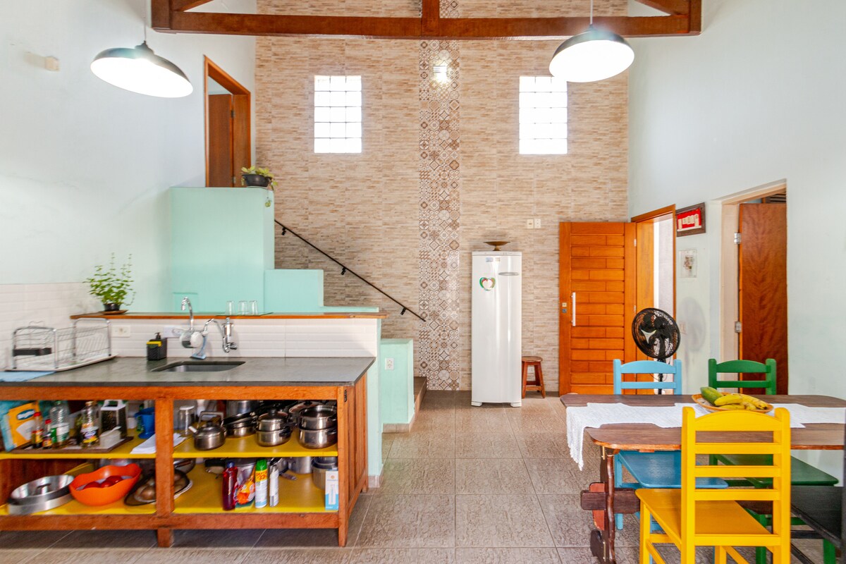 A bright kitchen area is shown, featuring a spacious counter with cooking utensils and a variety of pots. Colorful chairs surround a dining table, with a staircase leading to an upper level. Natural light enters through high windows, creating an inviting space.