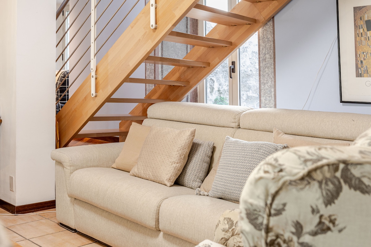 A light-toned sofa adorned with decorative cushions is positioned in a cozy seating area. A wooden staircase with a minimalist design leads to an upper level, while large windows illuminate the space with natural light.