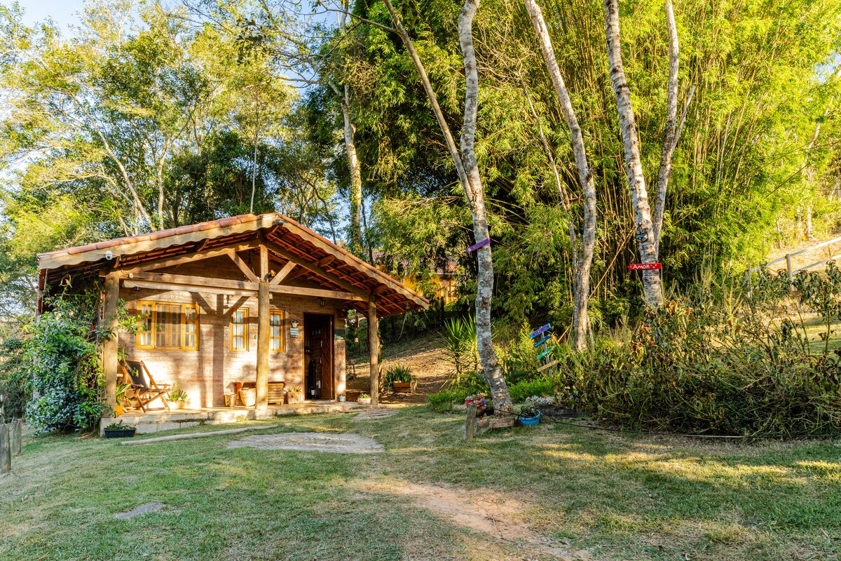 A charming chalé is surrounded by greenery, featuring a wooden entrance and a porch with seating. Lush grass and trees frame the structure, creating a peaceful atmosphere that promotes connection with nature.