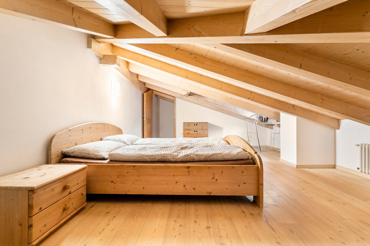 A spacious sleeping area features a wooden bed with a soft coverlet, complemented by wooden nightstands. The room is illuminated by natural light, highlighting the sloped wooden ceiling and light wooden flooring, creating a serene environment.