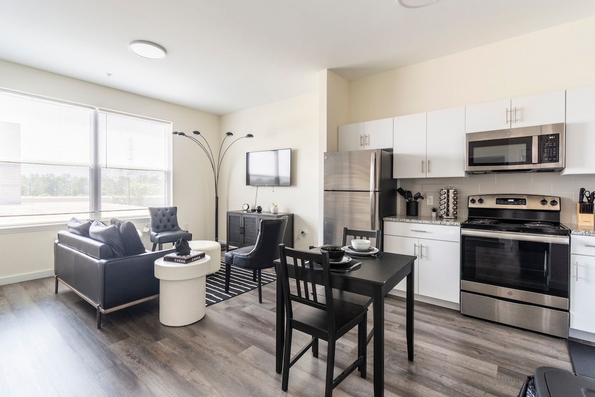 A spacious living area is highlighted by large windows allowing ample natural light. A sleek kitchen features modern appliances, while a comfortable seating arrangement includes a black armchair and a contemporary coffee table, enhancing the overall functional layout.