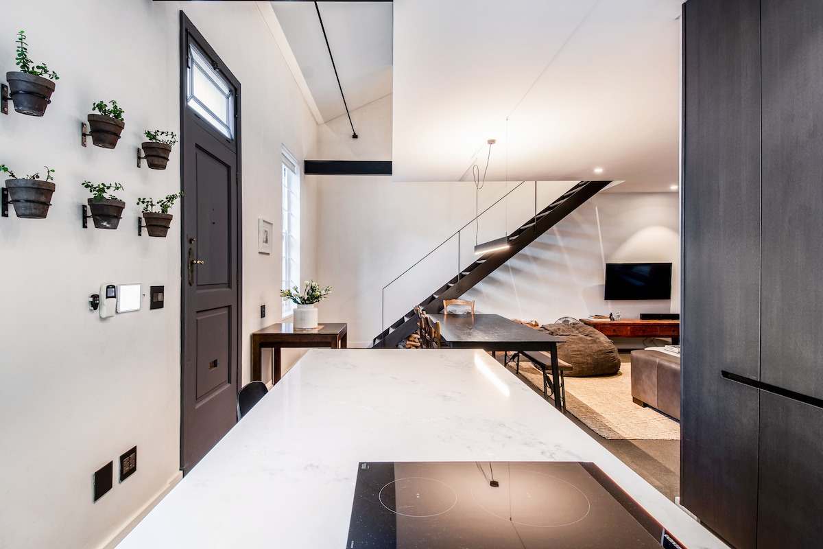 An open-plan living area is highlighted, showcasing a modern kitchen with a white countertop and integrated induction stove. A door with glass panels and a neutral wall creates a bright atmosphere. A staircase with black railings leads to the upper level, with a comfortable seating area visible in the background.