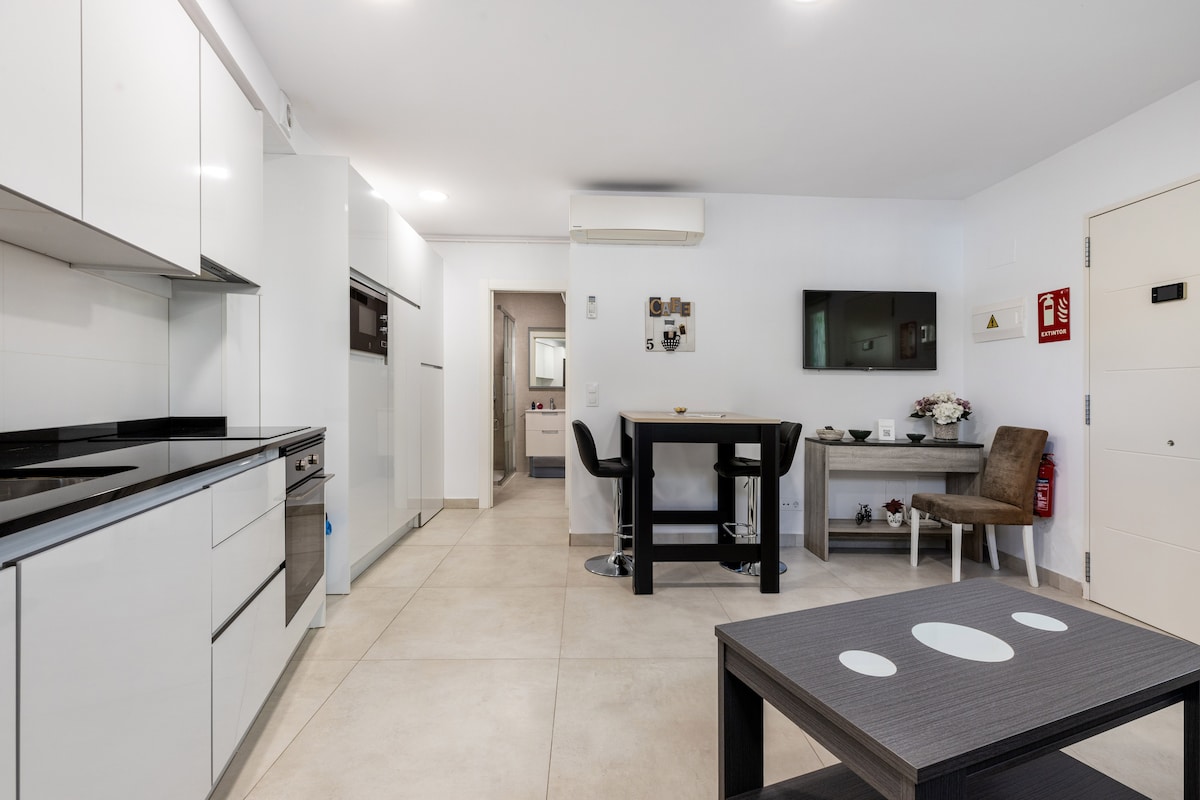 A modern kitchen is paired with a dining area in this open layout. Sleek, white cabinetry contrasts with dark countertops. A small table and four chairs invite casual dining. A wall-mounted TV is positioned in front of a cozy sitting area featuring light-colored furnishings.