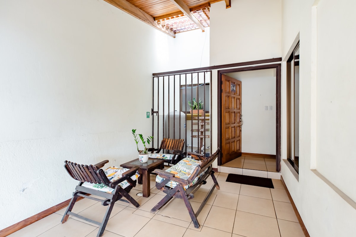 An entryway features two wooden chairs with cushions set around a small table. A door is visible in the background, leading to the rest of the apartment. Natural light enters through a window above, illuminating the tiled floor and white walls.