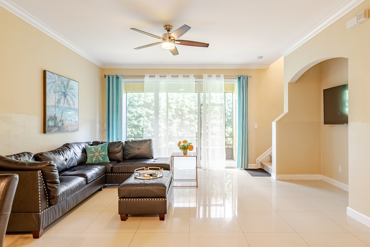 A spacious living area features a dark leather sectional sofa complemented by a decorative ottoman. Natural light filters through sheer curtains, illuminating the tiled floor. An accent wall displays a tropical-themed artwork, and a flat-screen television is mounted on a wall opposite the seating area.