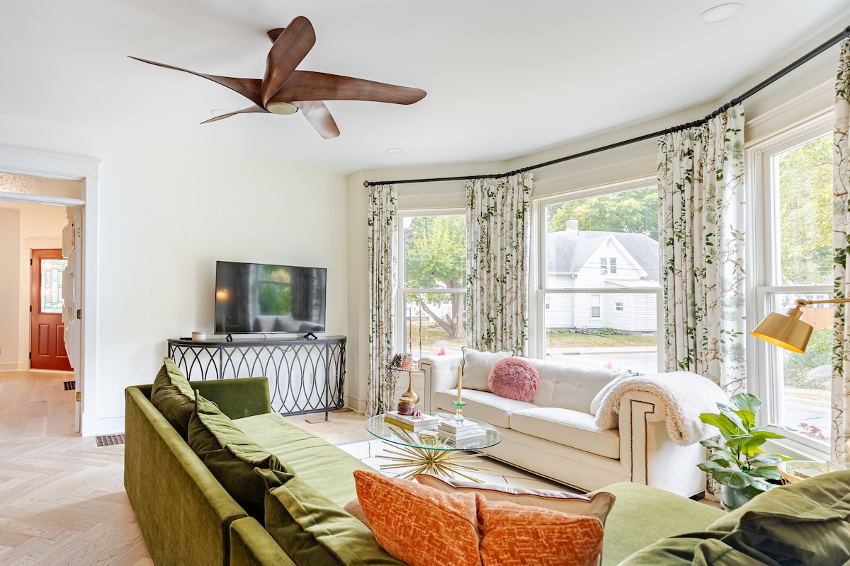 A bright living room features large windows that allow ample natural light to fill the space. A green sectional sofa is paired with a cozy cream-colored couch. A glass coffee table sits in the center, surrounded by stylish accents, and a ceiling fan enhances airflow.