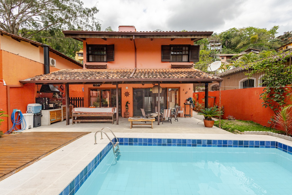 An outdoor area features a swimming pool with a light blue tiled edge. Adjacent to the pool, a covered patio is equipped with seating and a dining area. The house exhibits a bright exterior with earthy tones and terracotta roofing, surrounded by greenery.