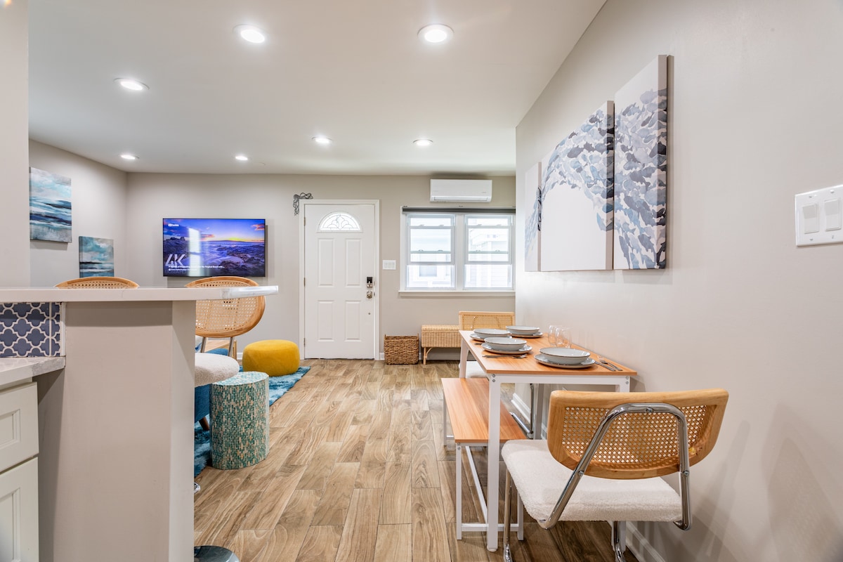 An open living area is presented, featuring a modern design with wood-look flooring. A flat-screen TV is mounted along the wall, while a dining table with two chairs occupies one corner. Natural light enters through a window near the entrance door, creating a bright atmosphere.
