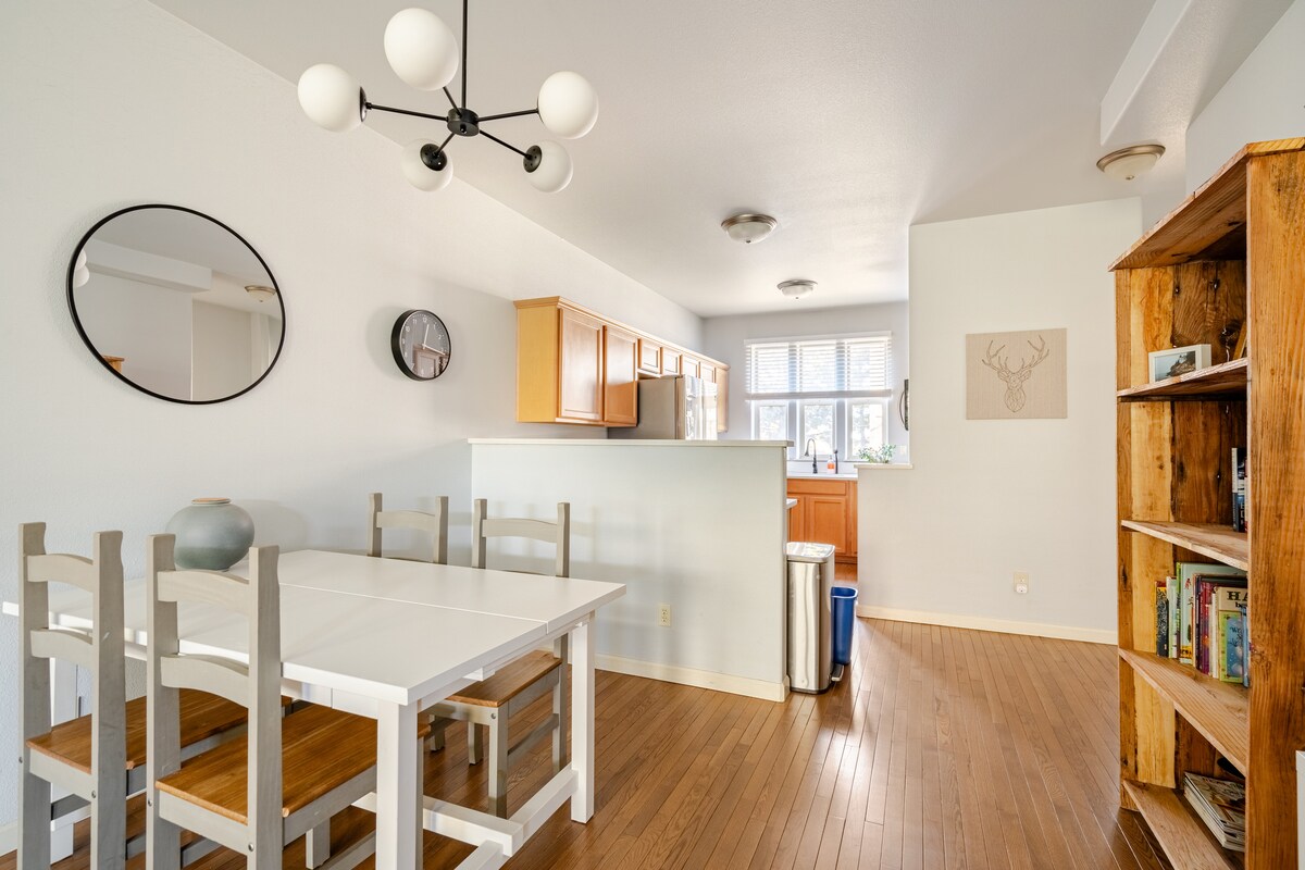 A dining area is shown with a white table and light wooden chairs, complemented by a circular mirror on the wall. Natural light filters in through multiple windows, illuminating the modern kitchen in the background and a wooden bookshelf filled with books.