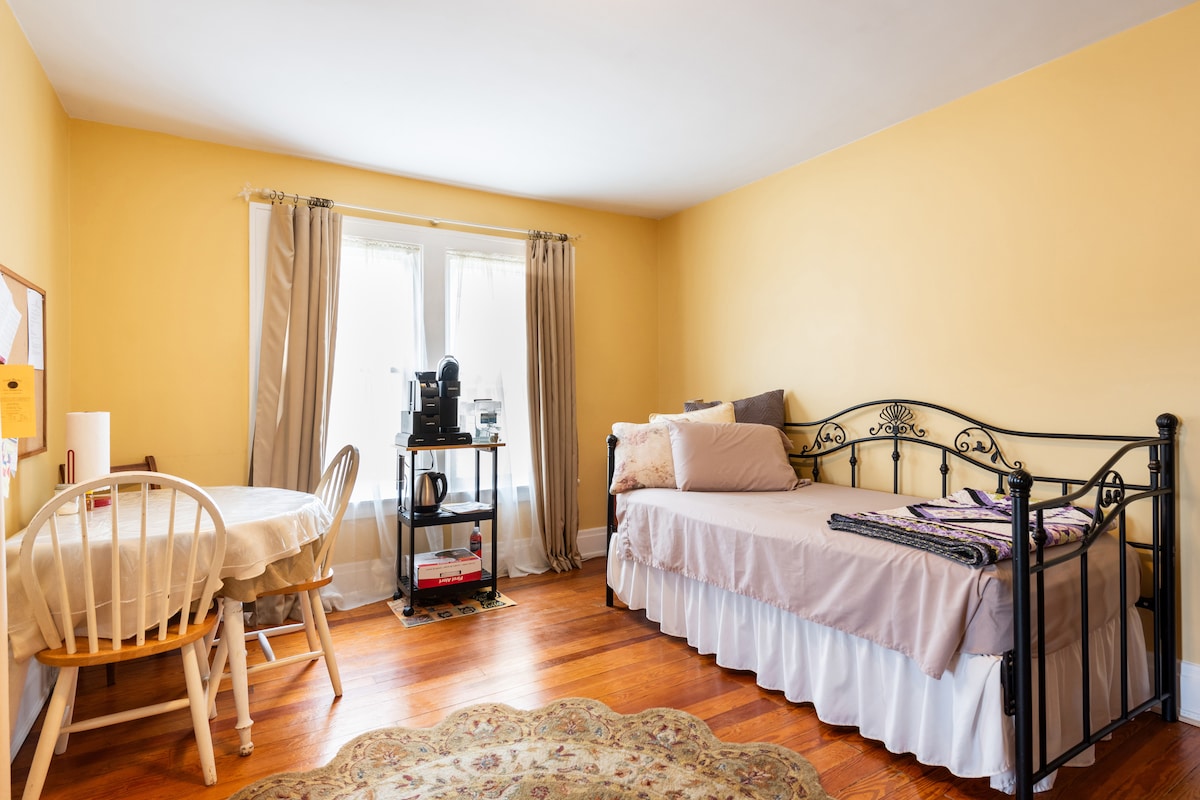 A bright, inviting room features a daybed with soft bedding and a round table with two chairs. Natural light pours through two windows, partially covered by curtains. A small coffee station is set up on a black cart, enhancing the space's functional appeal.