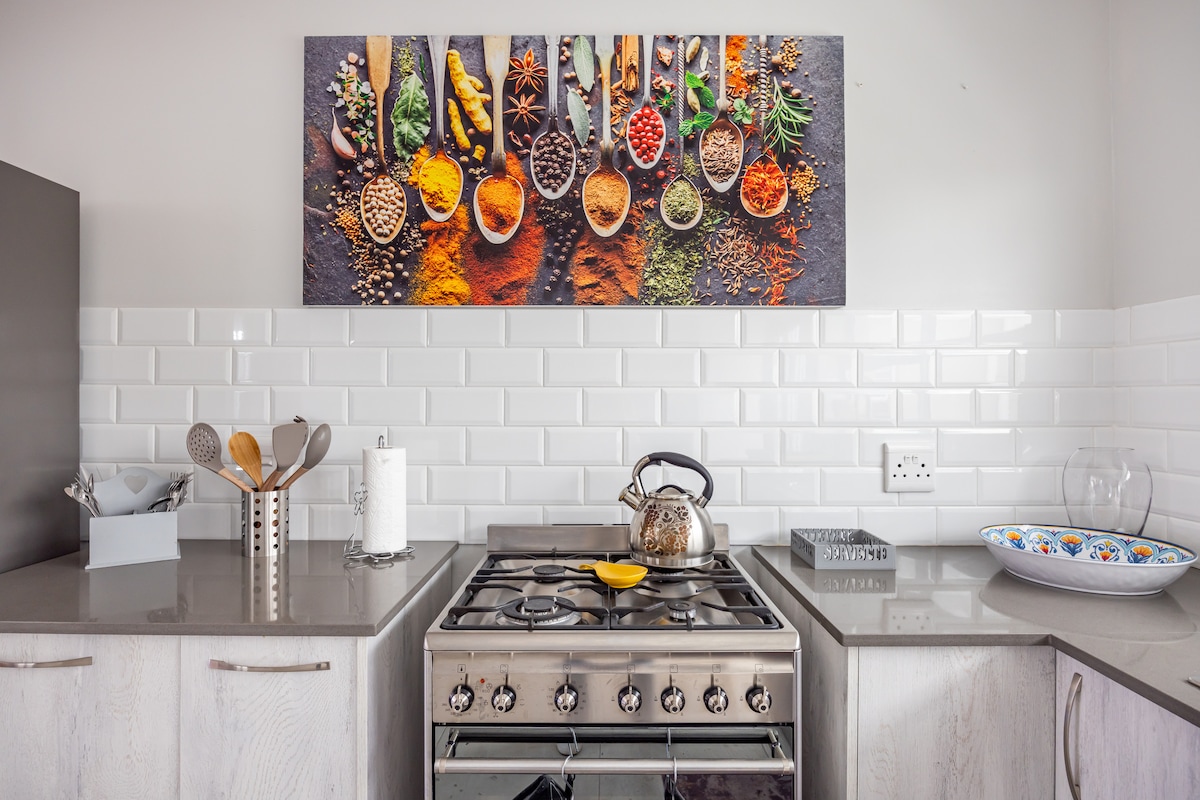 A modern kitchen is showcased with sleek cabinetry and a stainless-steel stove. Above, a vibrant wall art piece displays an array of spices and herbs on wooden spoons. A kettle is placed on the stove, complemented by organized utensils and a decorative bowl on the counter.