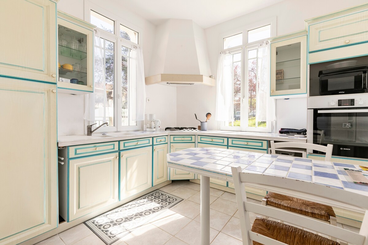 The kitchen features light-colored cabinetry with teal accents, complemented by large windows allowing natural light to fill the space. A central table with a blue and white patterned top is surrounded by wooden chairs. Modern appliances and a hood above the stove are also visible.