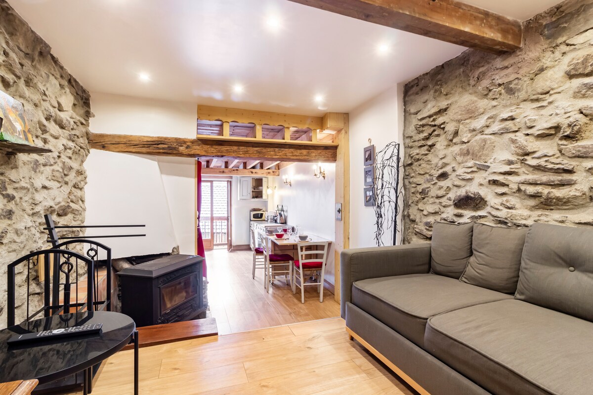 A cozy living area features a comfortable sofa adjacent to a wood stove. Exposed stone walls and wooden beams create a rustic ambiance. An open kitchen and dining area are visible in the background, with a dining table and chairs set against light-colored cabinetry.