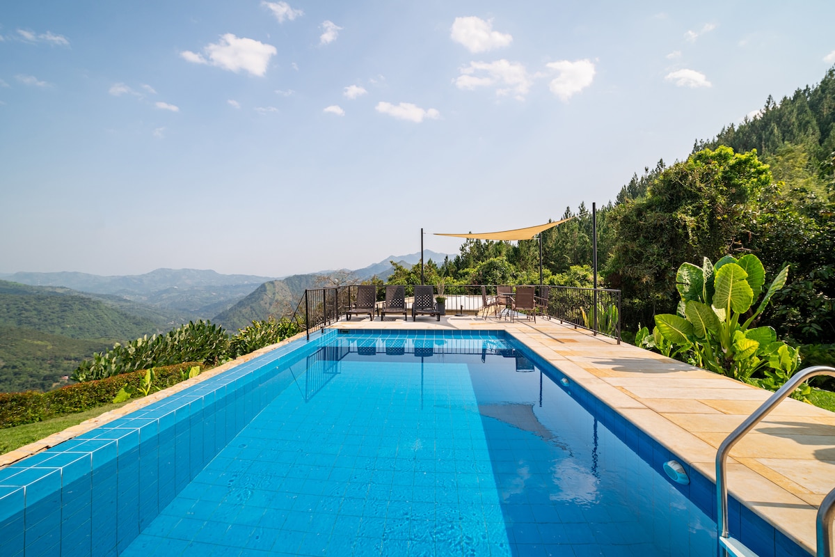 A tranquil swimming pool is set against a backdrop of rolling mountains, featuring clear blue water and a surrounding stone patio. Lounge chairs are arranged nearby, inviting relaxation under a shaded area. Lush greenery extends beyond the pool, contributing to the serene landscape.