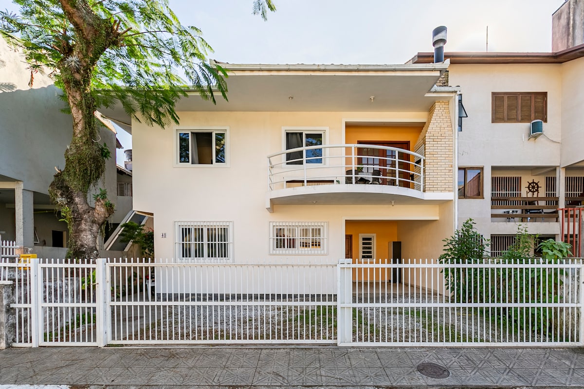 The exterior of the house showcases a white façade with large windows and a welcoming balcony. A white picket fence surrounds the property, and lush greenery is visible, creating a tranquil environment. The building's two levels are easily distinguished, highlighting its spacious layout.