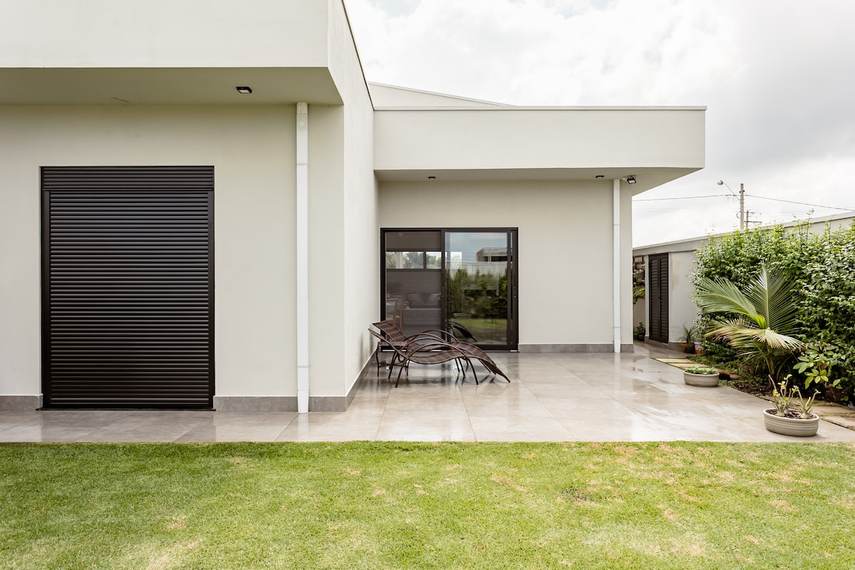 The exterior of a modern house is displayed, featuring clean lines and a combination of smooth wall surfaces. A spacious patio area is shown, with several chairs arranged around a small table, surrounded by greenery. Dark shutters cover a window, and a palm plant adds a natural touch.