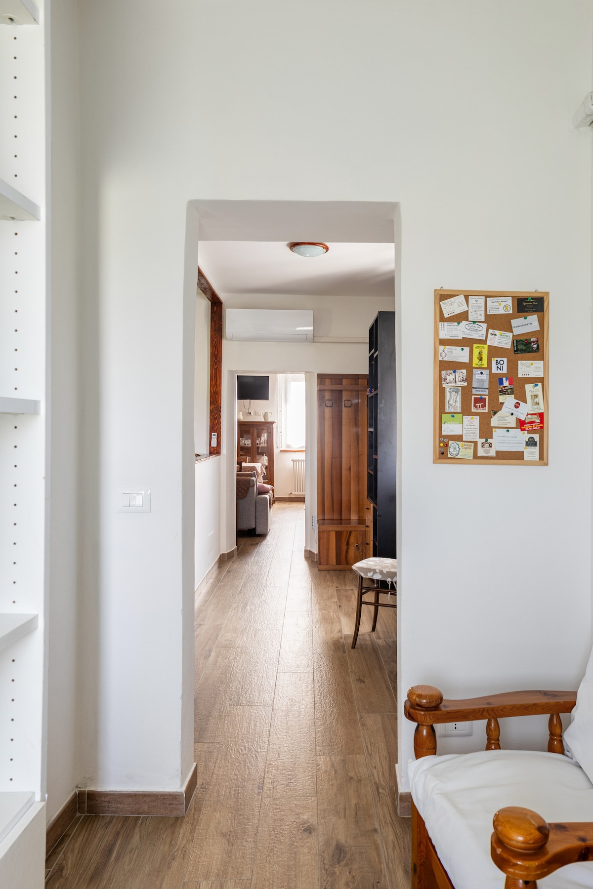 A well-lit hallway features a light wooden floor and white walls. On one side, a white shelf stands, while on the opposite side, a bulletin board displays various notes and cards. A glimpse of the living area can be seen at the end of the hall.