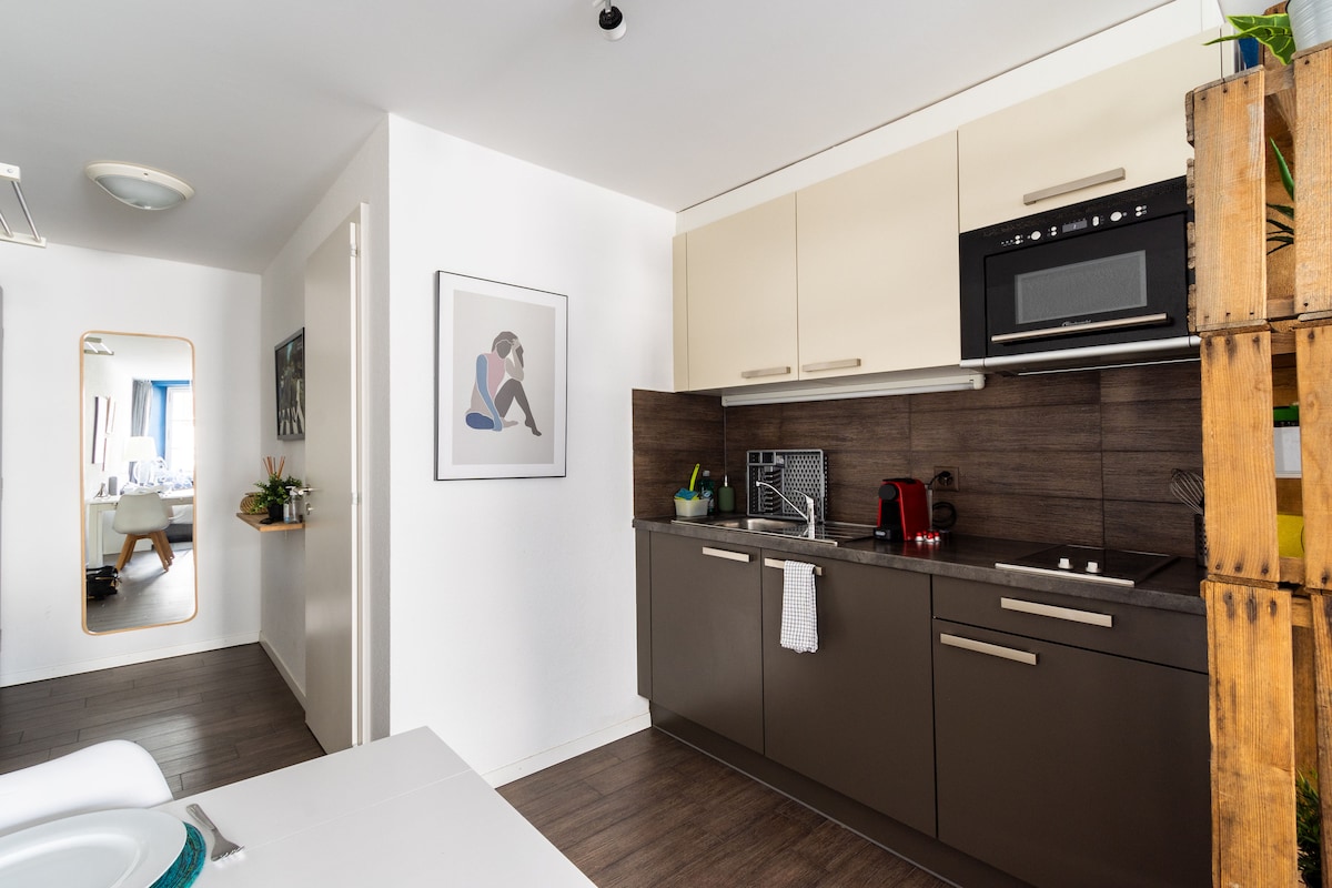 A modern kitchen area is designed with sleek cabinetry and dark countertops. A microwave and essential kitchen tools are visible. Natural light is filtered through a window, and a mirror reflects the adjacent space, enhancing the sense of openness.